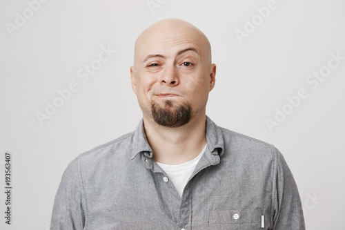 Close-up portrait of joyful attractive bald bearded man, looking suspicious or hesitation about something, standing against gray background. Guy is not sure that friend told him truth