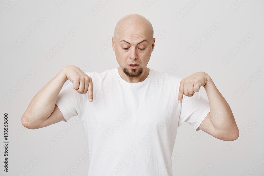 Portrait of intrigued bald european bearded man pointing down and looking at floor as if seeing something surprising, standing against gray background in white t-shirt. Guy looks from balcony Portrait of intrigued bald european bearded man pointing down and looking at floor as if seeing something surprising, standing against gray background in white t-shirt. Guy looks from balcony