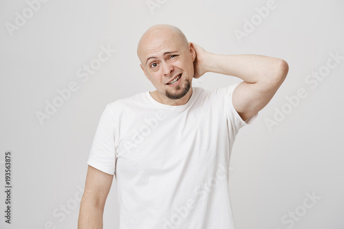 Obraz na plátně Portrait of bothered bald caucasian bearded male scratching neck while being doubtful, frowning and expressing confusion, standing against gray background