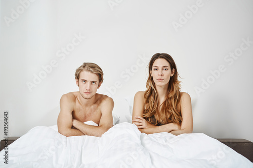 Bedroom shot of couple sitting in bed naked under blanket with crossed hands and unsatisfied expression. Young adults in relationship have problems in their sexual life they do not want discuss