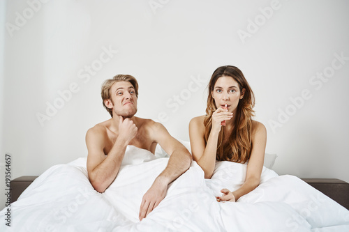 Attractive european wife sitting in bed with husband, applying lip balm while he feels boredom and thinks about something. Couple recently woke up and boyfriend remembers where is TV remote.