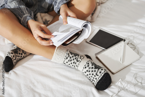 Side angle shot of woman sitting with crossed hands on bed, wearing fancy socks, making notes while studying at home. Student working on homework, using digital tablet, listening music with headphones
