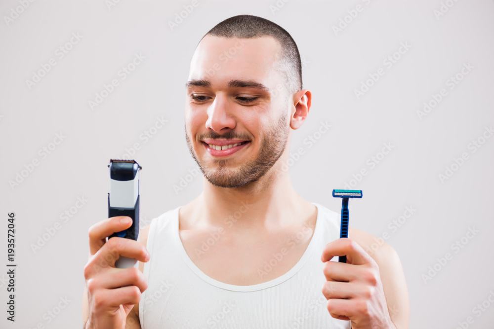 Young man is holding electric and classic razor. He is deciding which one to use to shave his beard. Young man is holding electric and classic razor. He is deciding which one to use to shave his beard.