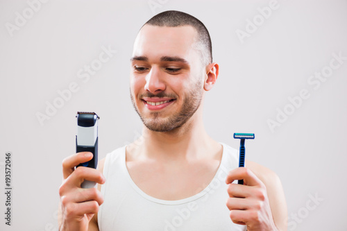Young man is holding electric and classic razor. He is deciding which one to use to shave his beard. 