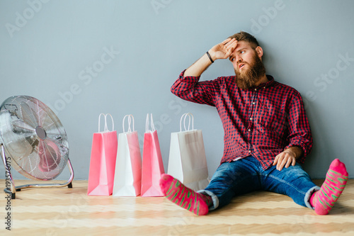 Full length portrait of a hipster tired chill beard man in colorful socks, jeans and shirt sitting on the floor with pink and white shopping bags under gray wall background. Facial expressions concept photo