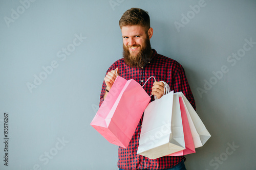 Canvas Print Playful hipster beard man in casual clothes holding pink shopping paper bags, sitting on the floor under gray wall background