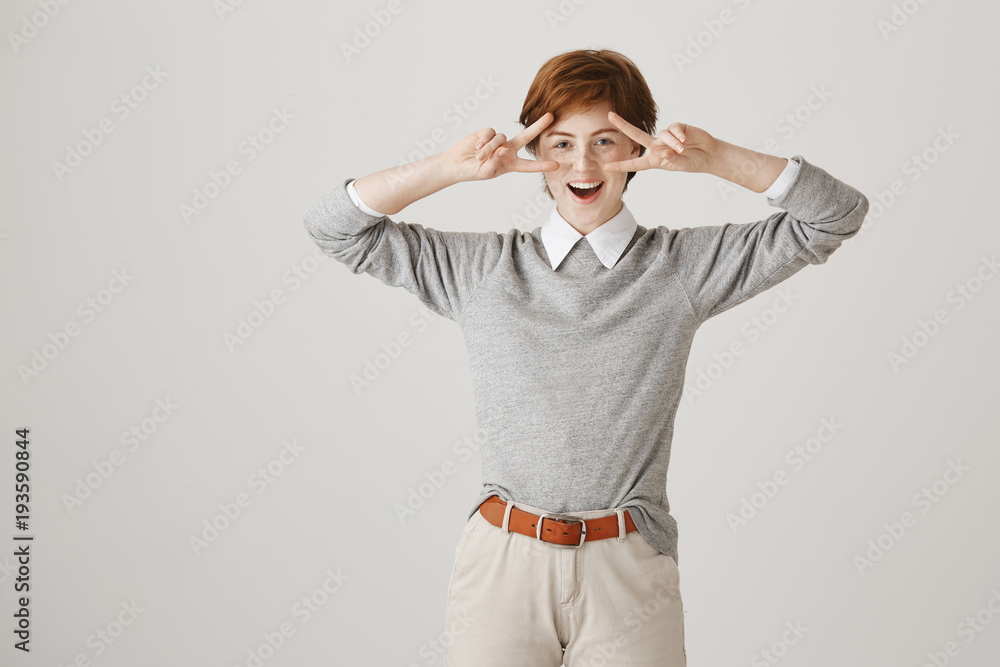 Playful excited european girlfriend with short haircut and freckles standing in boyish clothes and showing victory or peace gestures over eyes, smiling broadly and expressing positive emotions. Playful excited european girlfriend with short haircut and freckles standing in boyish clothes and showing victory or peace gestures over eyes, smiling broadly and expressing positive emotions.