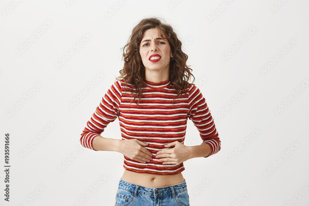 My stomach aches so bad. Studio shot of attractive female in cropped top touching belly with hands while feeling pain or discomfort, expressing negative emotions with mimic, frowning over gray wall My stomach aches so bad. Studio shot of attractive female in cropped top touching belly with hands while feeling pain or discomfort, expressing negative emotions with mimic, frowning over gray wall