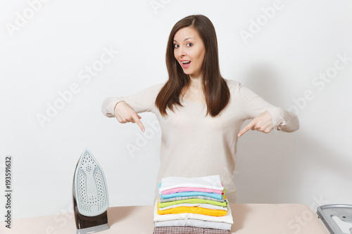 Young fun housewife in light clothes pointing index fingers on colorful family clothing on ironing board with iron. Woman isolated on white background. Housekeeping concept. Copy space advertisement.
