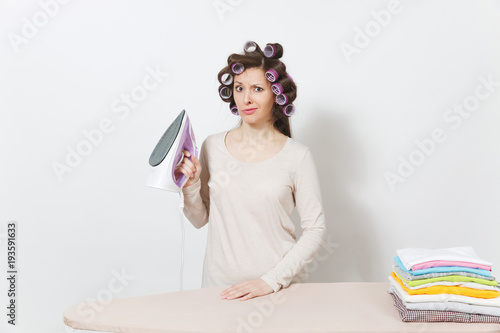 Crazy fun housewife with curlers on hair in light clothes ironing family clothing on ironing board with iron. Woman isolated on white background. Housekeeping concept. Copy space for advertisement.