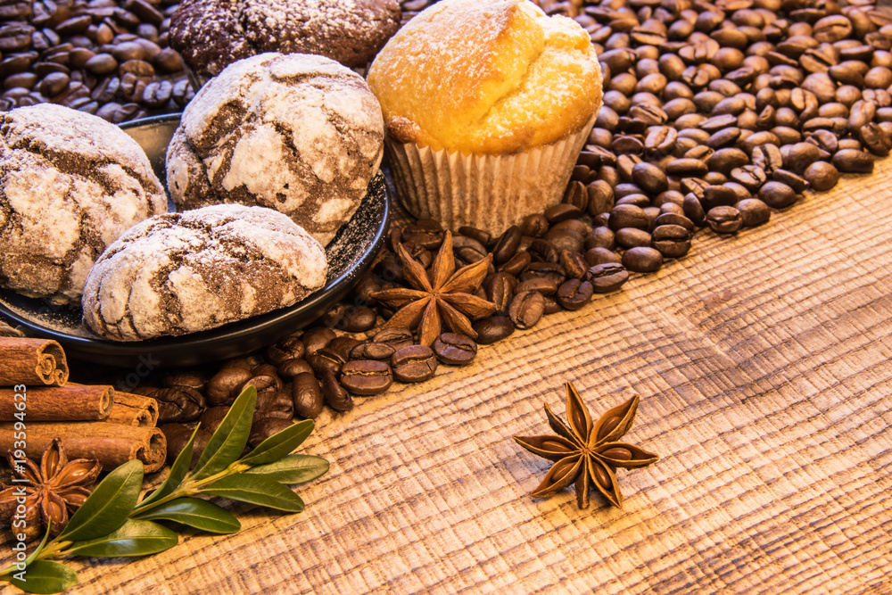 On a wooden table there is a mug of black coffee next to a plate of chocolate cookies sprinkled with powdered sugar, and on the background coffee beans spread with cinnamon and puddings