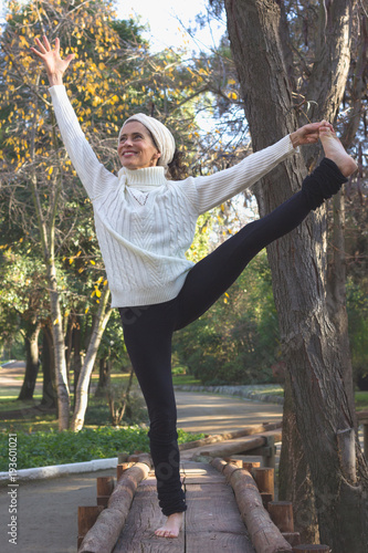 Smiling middle aged woman in yoga standing balance Parsva Utthita Hasta Padangusthasana pose, known as extended hand to big toe pose, on wooden path in the park early morning during fall season