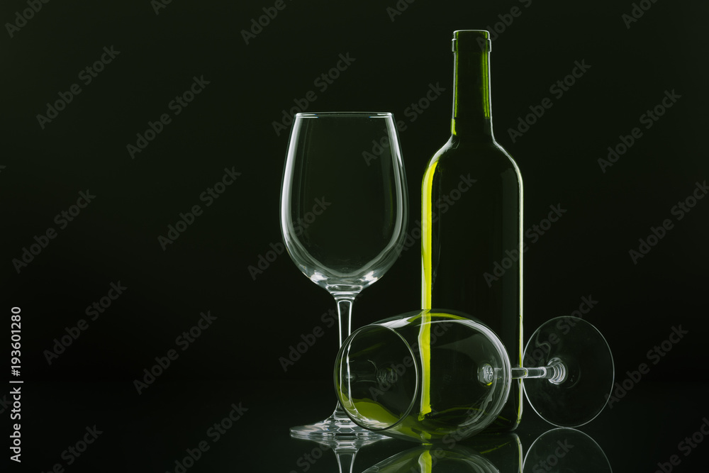 two wine glasses, one of them lying down and bottle of wine with green reflections with black background