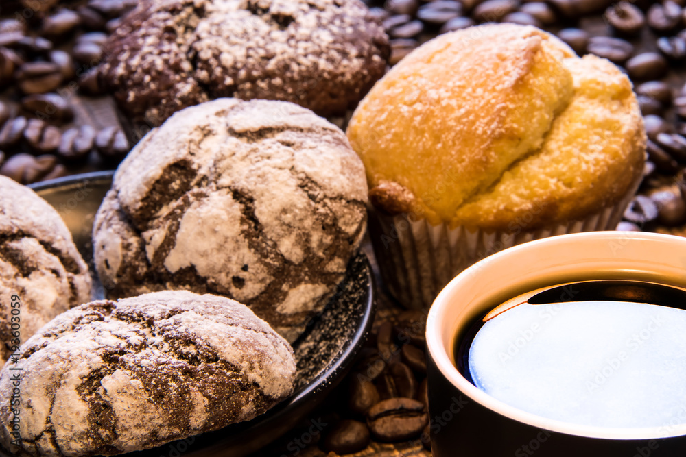 Close up mug of black coffee next to a plate of chocolate cookies sprinkled with powdered sugar, and on the background coffee beans