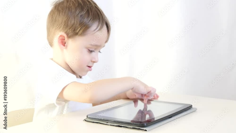 Happy child boy sitting at table, playing with tablet. The child is happy to play electronic games. The concept of child development. Motion camera slider. Happy child boy sitting at table, playing with tablet. The child is happy to play electronic games. The concept of child development. Motion camera slider.