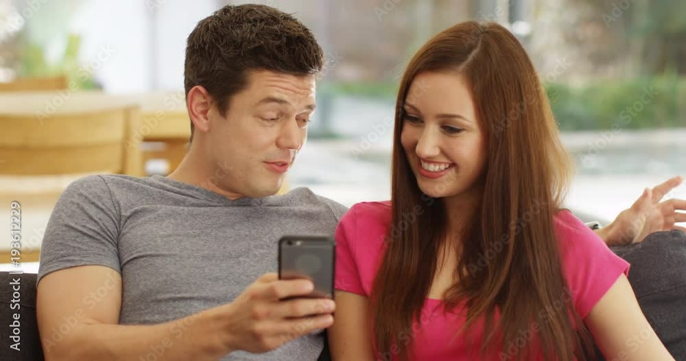 4K Happy young couple relaxing at home, talking & looking at smartphone 4K Happy young couple relaxing at home, talking & looking at smartphone