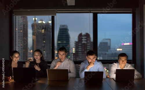 Business people team working late night in low light from laptop screen with cityscape blurred background.Overtime hours concept.
