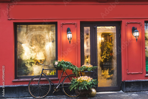 Christmas decorated showcase with old bicycle, fir branches, glass shiny toys and vintage lantern. Red building facade