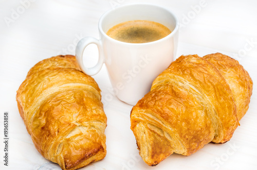 Two croissants and cup of coffee on the white table.