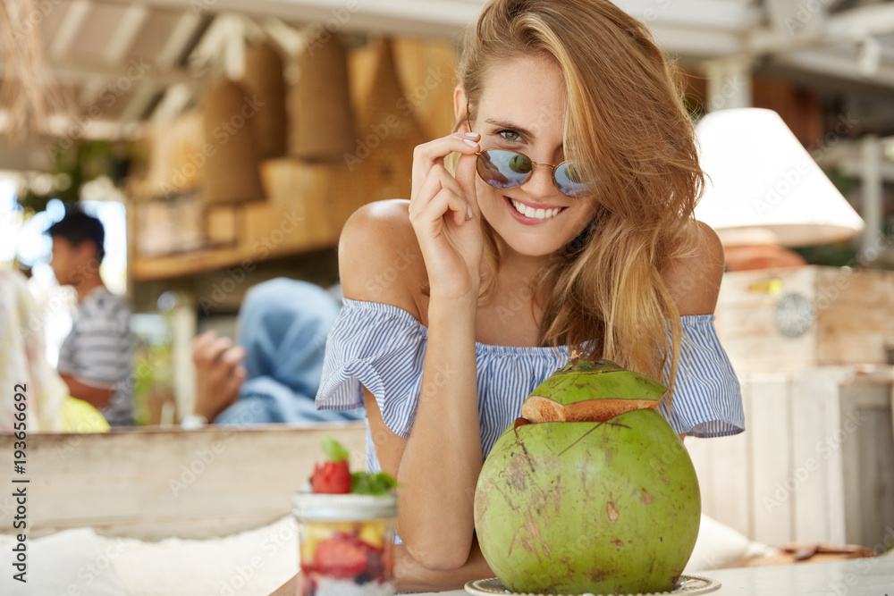Pleasant looking young female wears sunglasses, blouse, drinks coconut cocktail, sits against cafe interor, dressed in fashionable clothing, smiles pleasantly. Happiness and leisure concept.