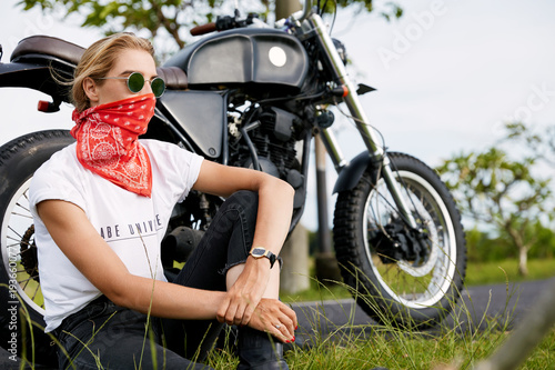 Stylish biker model poses near motorbike, sit on ground outdoor, wears sunglasses and bandana on face, feels freedom of motocycling riding. People, active lifestyle and transportation concept photo