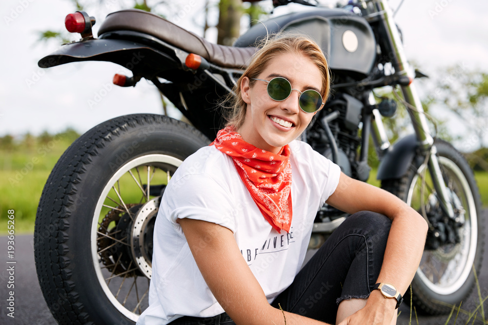 Cheerful relaxed female driver parks on road, poses against motorbike, sits on ground, has fun during driving, enjoys long route across beautiful landscapes outdoor. Travelling across world. Cheerful relaxed female driver parks on road, poses against motorbike, sits on ground, has fun during driving, enjoys long route across beautiful landscapes outdoor. Travelling across world.