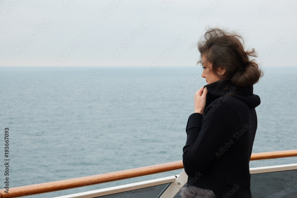 The girl is standing on the deck of a cruise liner and looks out into the ocean in windy autumn weather