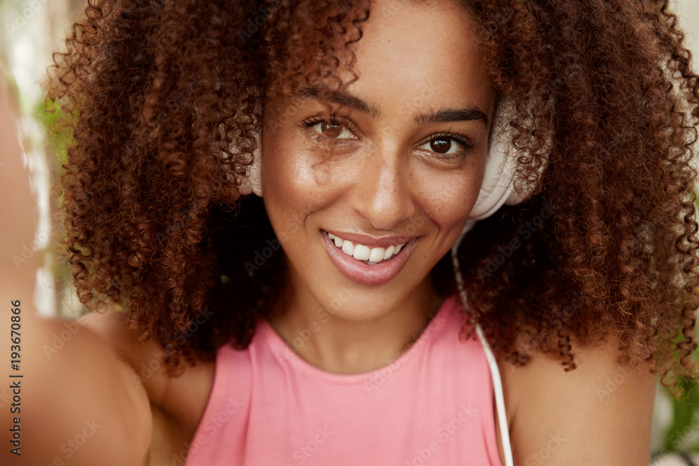 Attractive dark skinned female with curly bushy dark hair, wears headphones, listens audio book, shows pleasant smile, makes selfie, enjoys spare time. Student rests after university lessons Attractive dark skinned female with curly bushy dark hair, wears headphones, listens audio book, shows pleasant smile, makes selfie, enjoys spare time. Student rests after university lessons