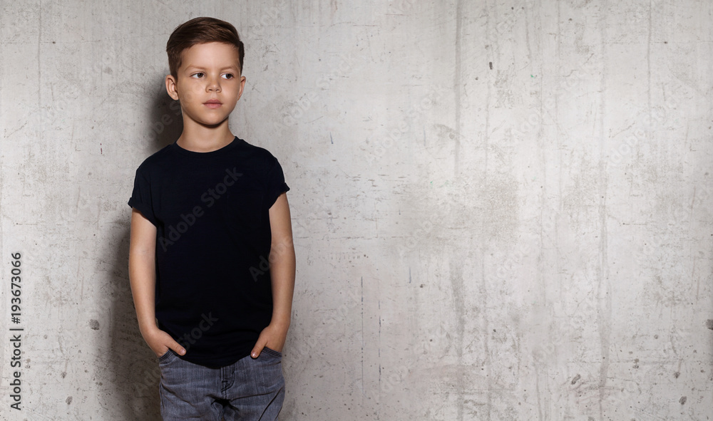 Wistful little guy stands on the wall background and looks away. Portrait of boy in black T-shirt, copy space. Concept of children style and fashion. Wistful little guy stands on the wall background and looks away. Portrait of boy in black T-shirt, copy space. Concept of children style and fashion.