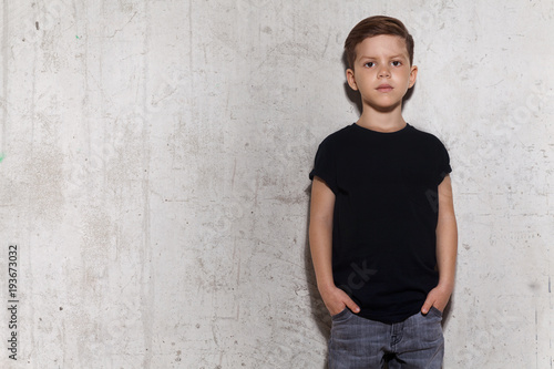 Cute little boy in black T-shirt posing in front of grunge concrete wall. Portrait of fashionable male child, copy space. Boy looks at camera, gray wall on background.