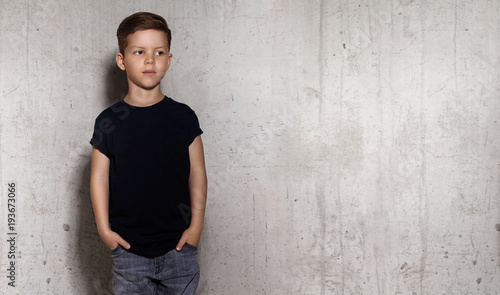 Wistful little guy stands on the wall background and looks away. Portrait of boy in black T-shirt, copy space. Concept of children style and fashion.