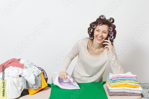 Young pretty housewife in light clothes, curlers on hair, talking on mobile phone, ironing family clothing on ironing board with iron. Woman isolated on white background. Copy space for advertisement.