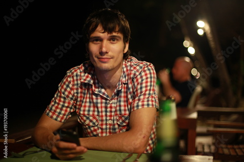 Young handsome man with smartphone drinking beer in beach bar sitting by sea at night with beautiful bokeh lights.