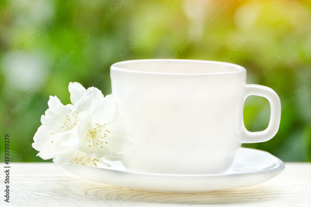 White mug of tea with jasmine on a background greenery, sunlight. Close up