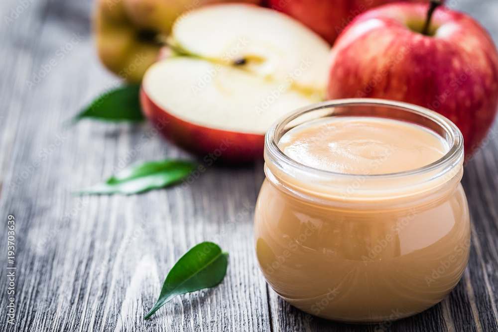 Homemade cinnamon canned applesauce in glass jar and red ripe apples on rustic wooden background. Selective focus, space for text. Homemade cinnamon canned applesauce in glass jar and red ripe apples on rustic wooden background. Selective focus, space for text.