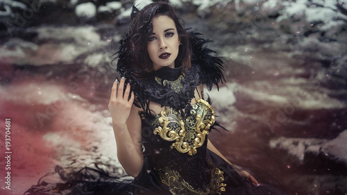Beautiful brunette woman with gothic dress made in gold and black threads. It is in a snowy forest in winter. fantasy concept