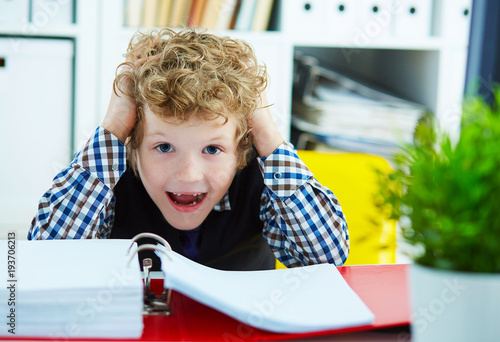 Small Caucasian curly boy grabbed his head while sitting over documents. Boy playing in the boss.