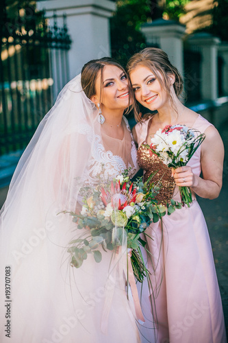 Wedding ceremony. Young bride and her bridesmaid hugging in beautiful summer park.