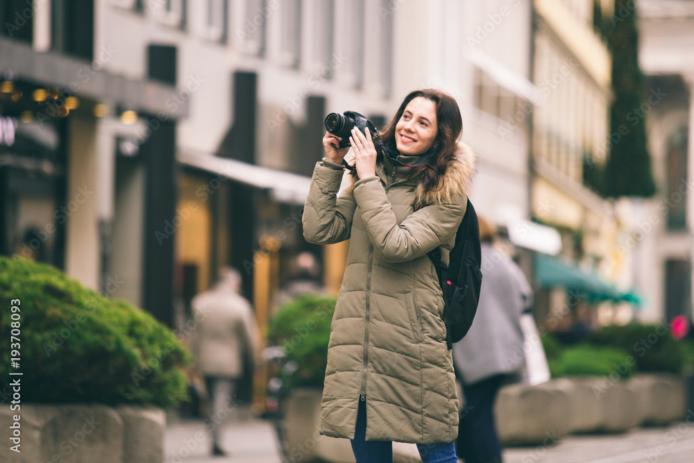 Beautiful young tourist woman stands on the background of the central street in Munich in Germany in winter. Holds a black big professional camera, takes a photo and smiles.