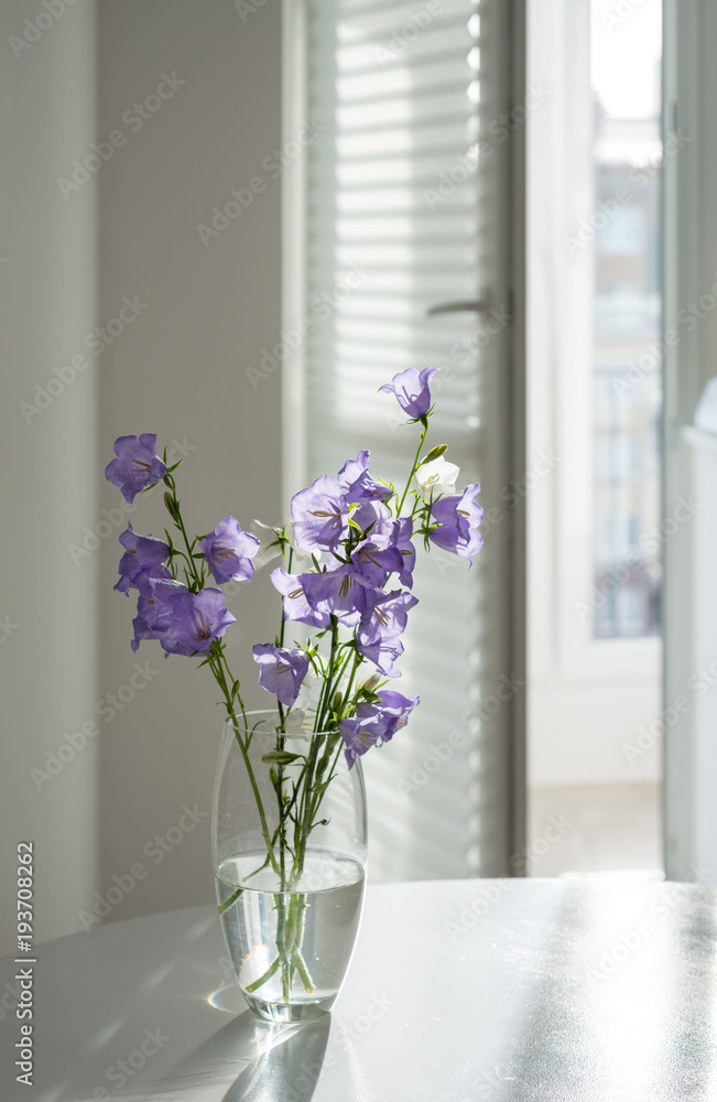 Flowers lilac bells in a glass vase stand on a white table on a window background
