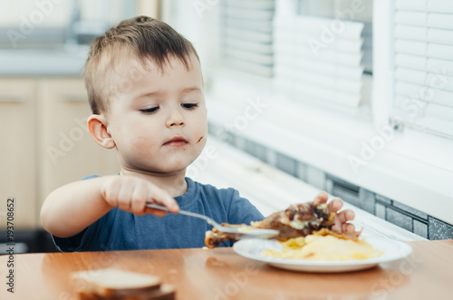 child in the kitchen eating mashed potatoes, spoon, next to the plate is a piece of chicken meat