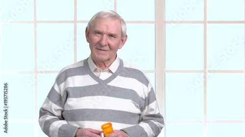 European elderly man with bottle of pills. Senior man holding container with medicaton and looking at camera, window background. photo