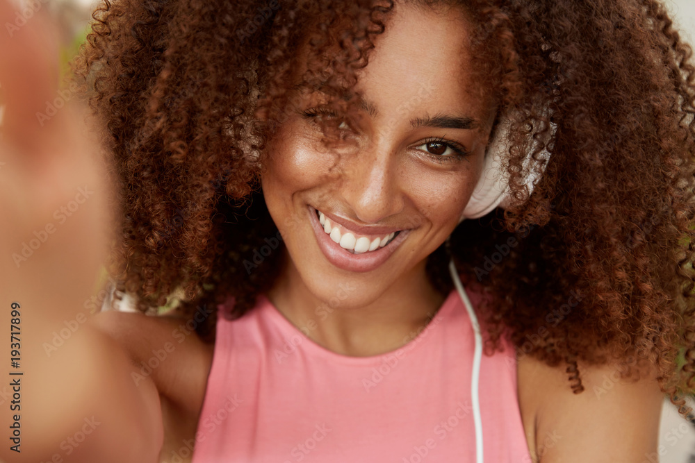 Portrait of curly beautiful African American woman in headphones, enjoys favourite music, makes photo of herself, has broad smile, dressed casually. Young dark skinned hipster girl poses for selfie Portrait of curly beautiful African American woman in headphones, enjoys favourite music, makes photo of herself, has broad smile, dressed casually. Young dark skinned hipster girl poses for selfie