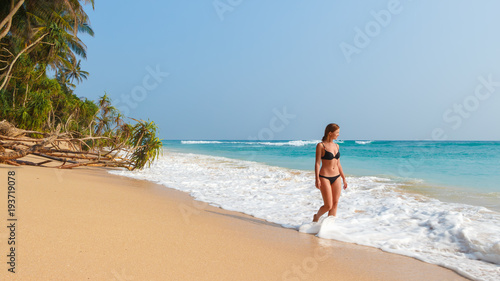 Young woman in black bikini on sand beach with palms near turquoise sea. Beautiful ocean view.