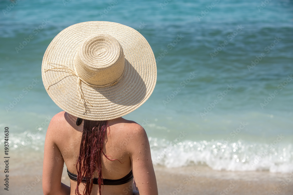The girl on the beach in a wide-brimmed hat sits and looks at the waves