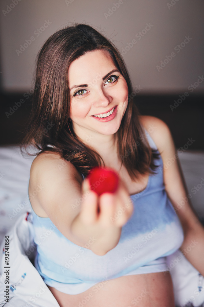 Attractive young pregnant woman sits on a bed and eats a ripe
