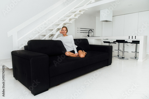 Attractive cheerful girl sitting on couch, posing, looking at camera. Modern design interior in black and white colors. At home.