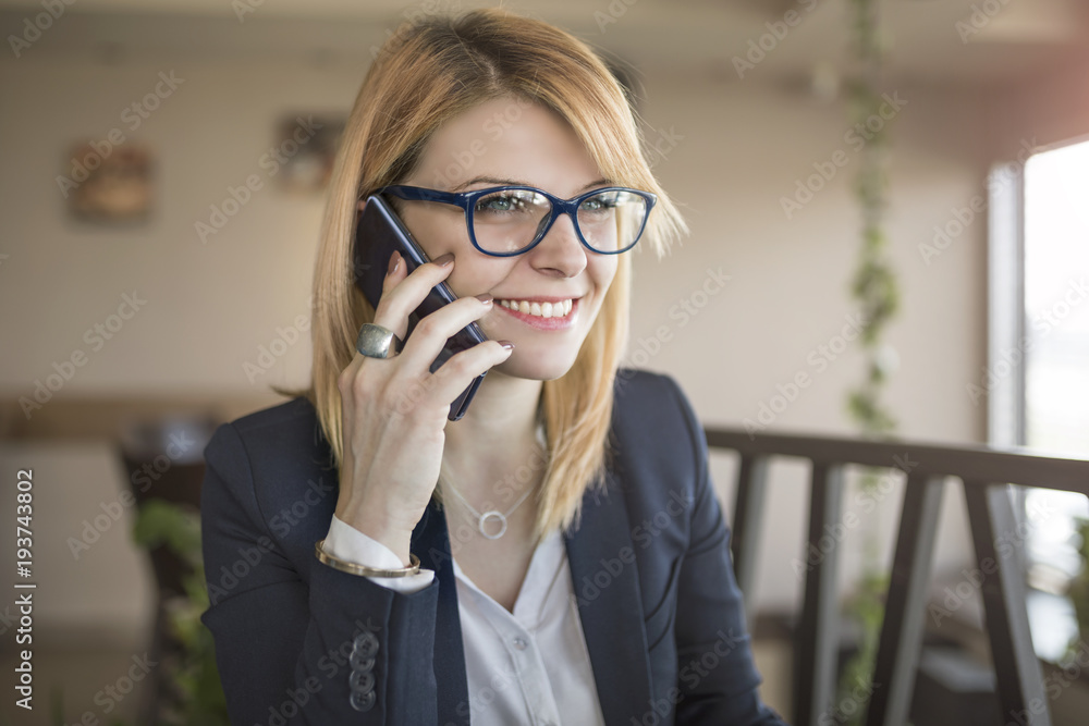 Cheerful young beautiful woman in glasses talking on mobile phone with smile while sitting in coffee shop, restaurant Cheerful young beautiful woman in glasses talking on mobile phone with smile while sitting in coffee shop, restaurant