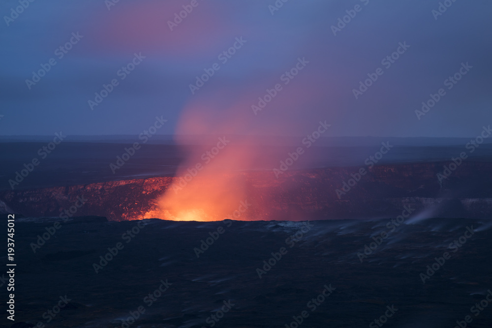 view of the kilauea crater at night showing the steam and smoke rising and the glow of the lava lake against the night sky and clouds with stars showing