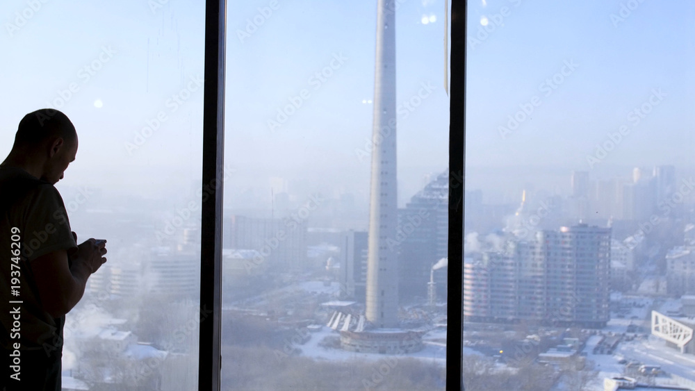 Fototapeta premium Silhouette of young man using smart phone with city view through glass windows. business and people concept - silhouette of businessman with smartphone over office window